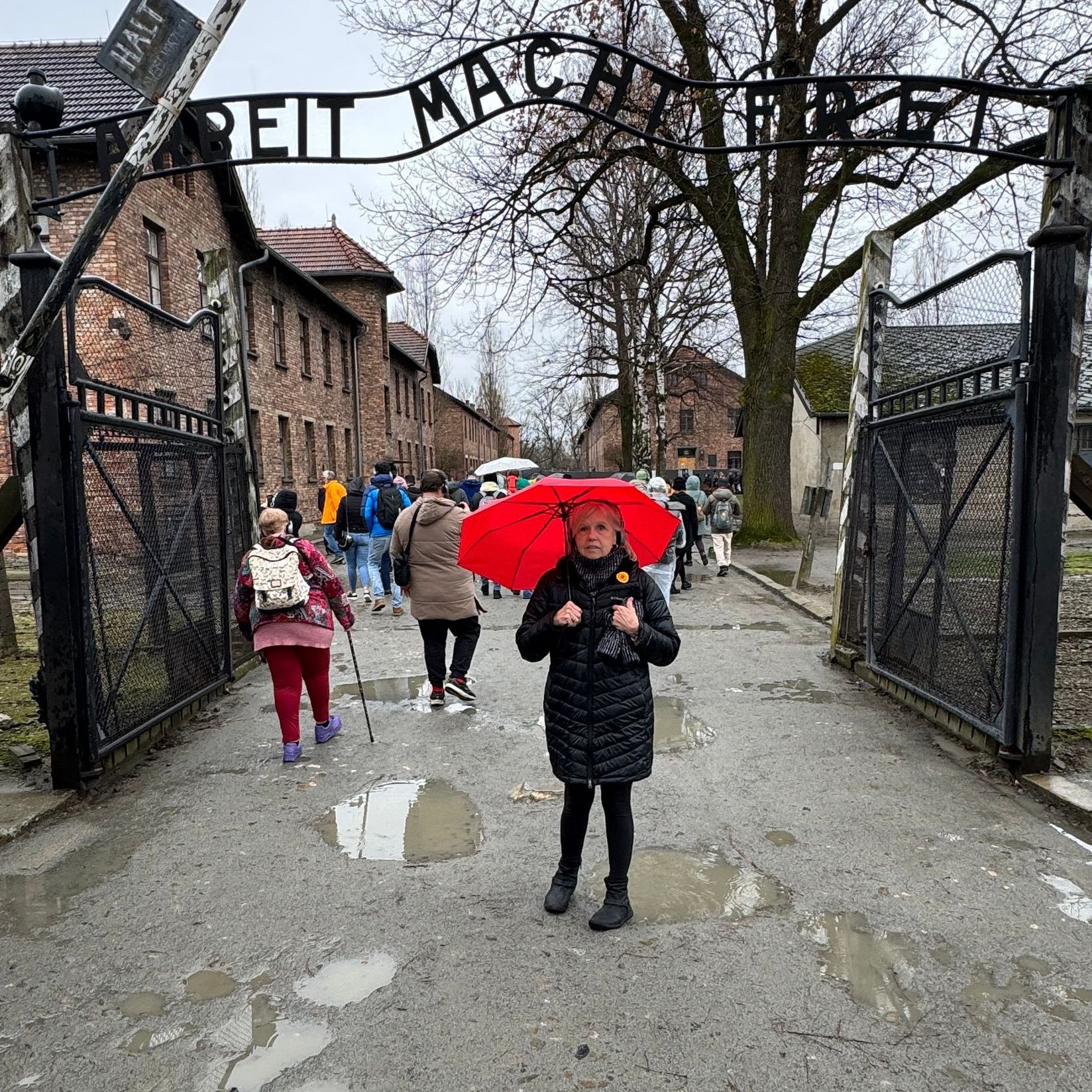 Teana holding an umbrella under a gray sky, standing beneath the gate into Auschwitz