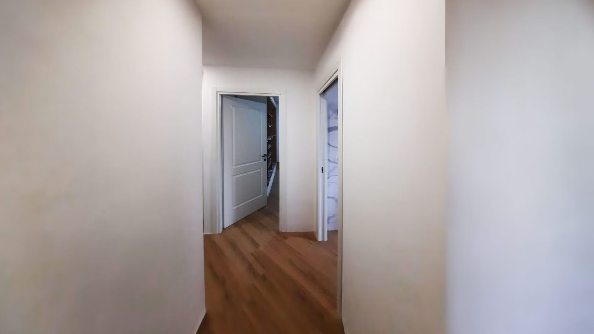 Un lungo corridoio con pavimento in legno e pareti bianche che conduce a una porta.
