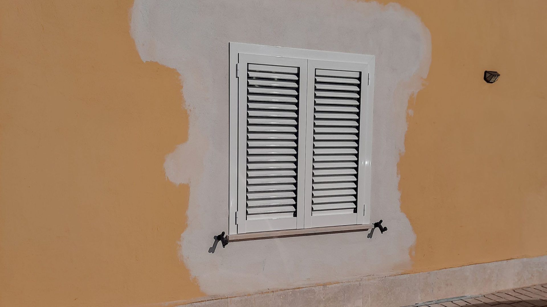 Una finestra bianca con persiane nere si trova su un muro marrone chiaro.