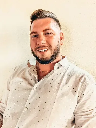 Un uomo con la barba indossa una camicia bianca e sorride alla telecamera.