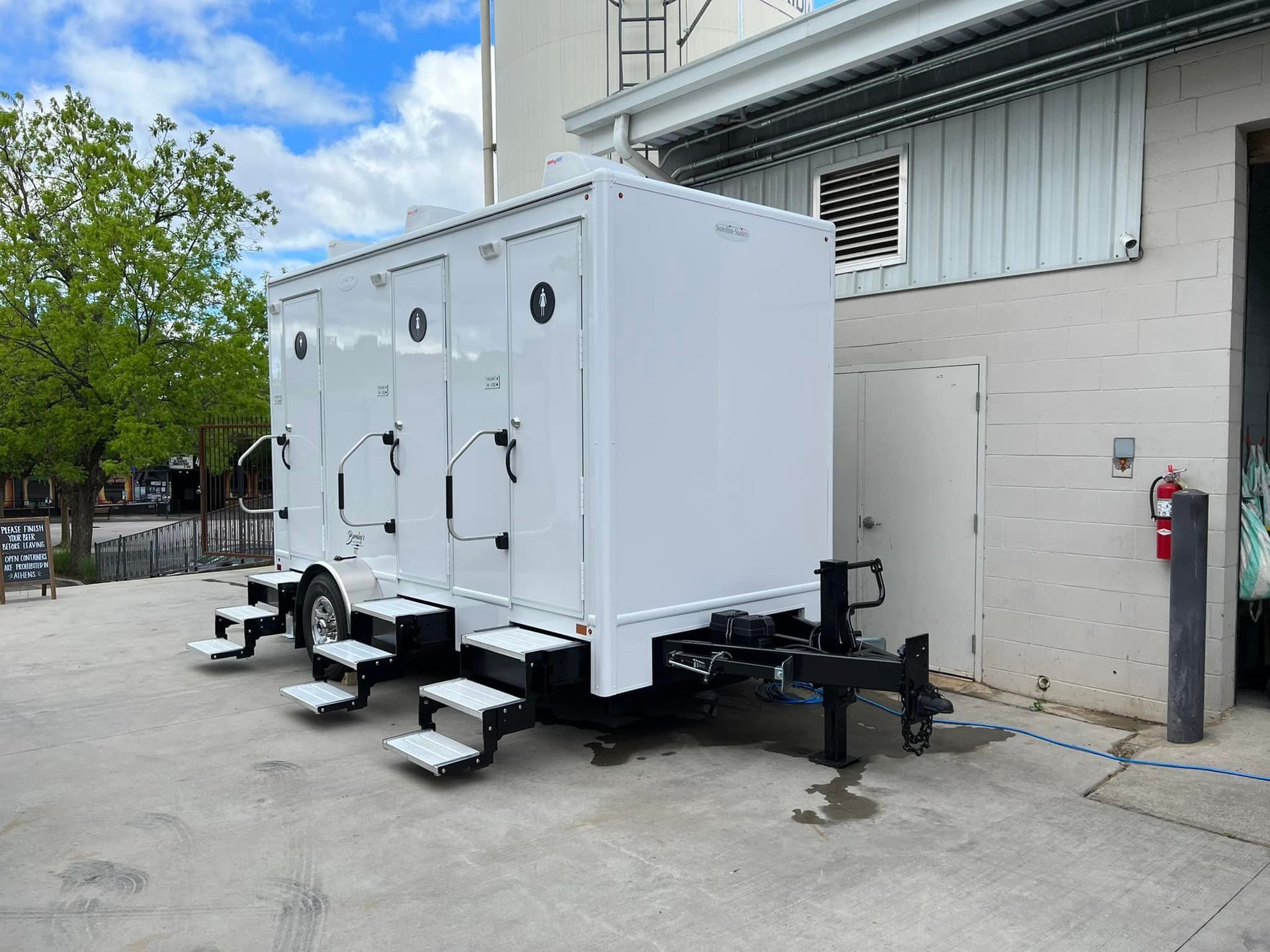 A white portable restroom trailer placed in a public space, featuring two white stairs and doors.