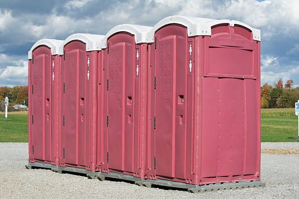 A row of maroon portable toilets with white tops on a gravel surface, outdoors.