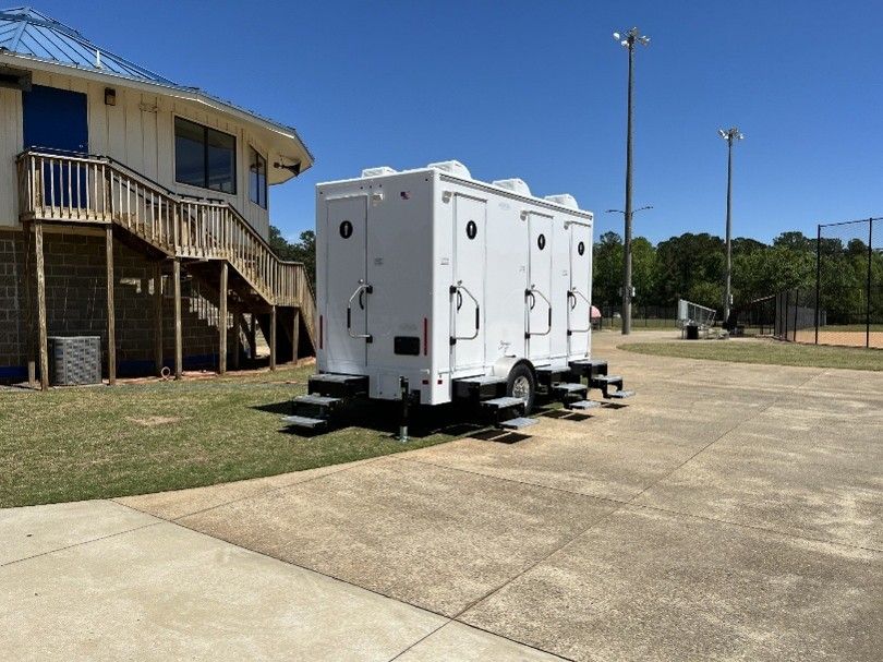 White portable restroom trailer on a concrete pad beside a building and grassy field under a sunny sky White portable restroom trailer on a concrete pad beside a building and grassy field under a sunny sky