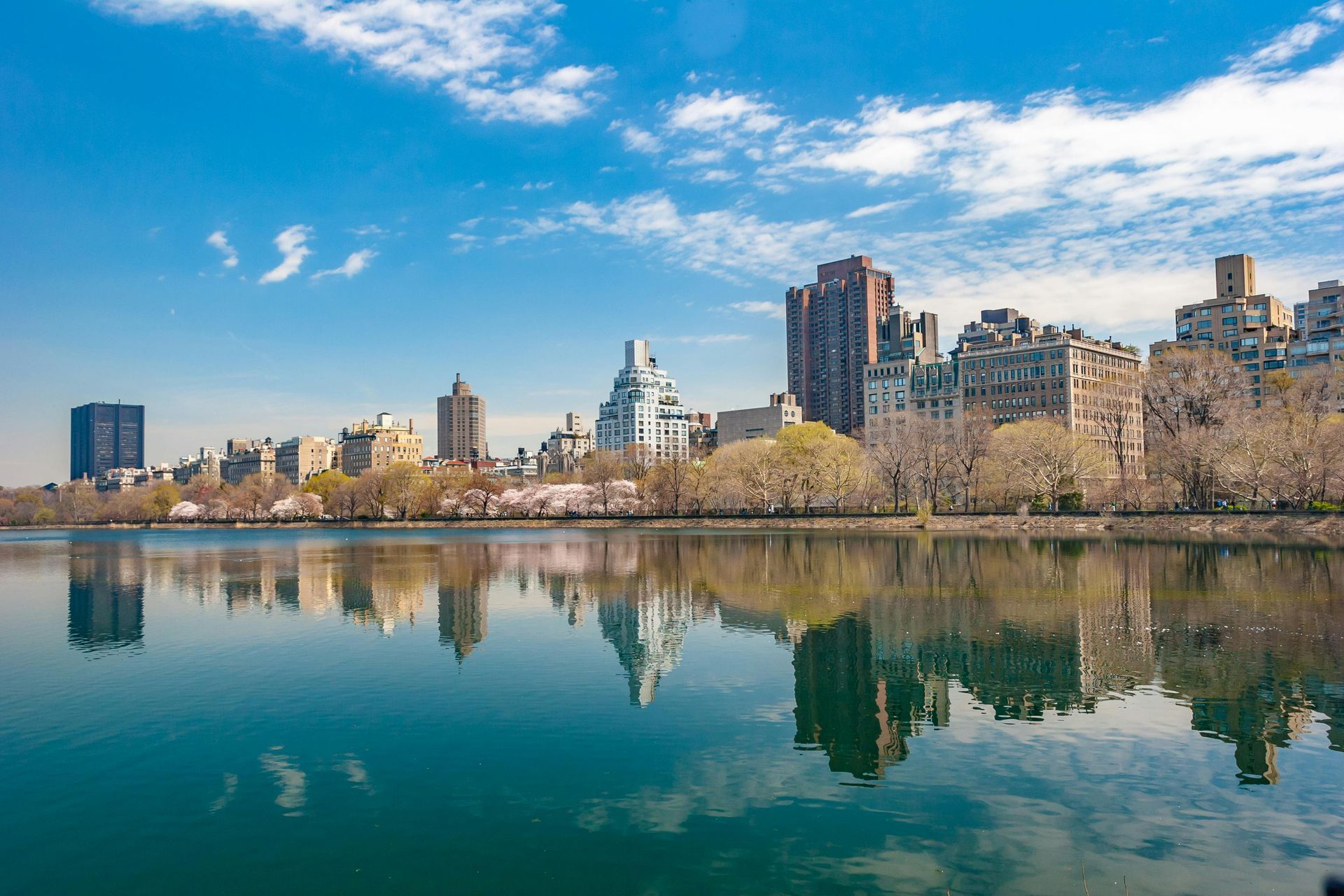A calm lake reflects a cityscape with blue sky and clouds.