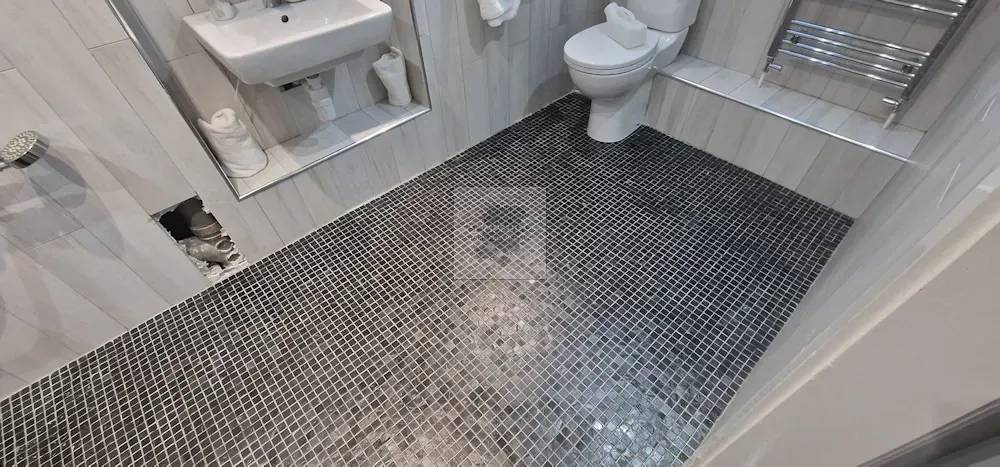 Bathroom interior, featuring a toilet, sink, and textured black and gray floor tiles. Damaged wall shows exposed structure.