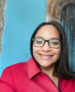 Woman with dark hair and glasses wearing a red jacket smiles in front of a blue wall.