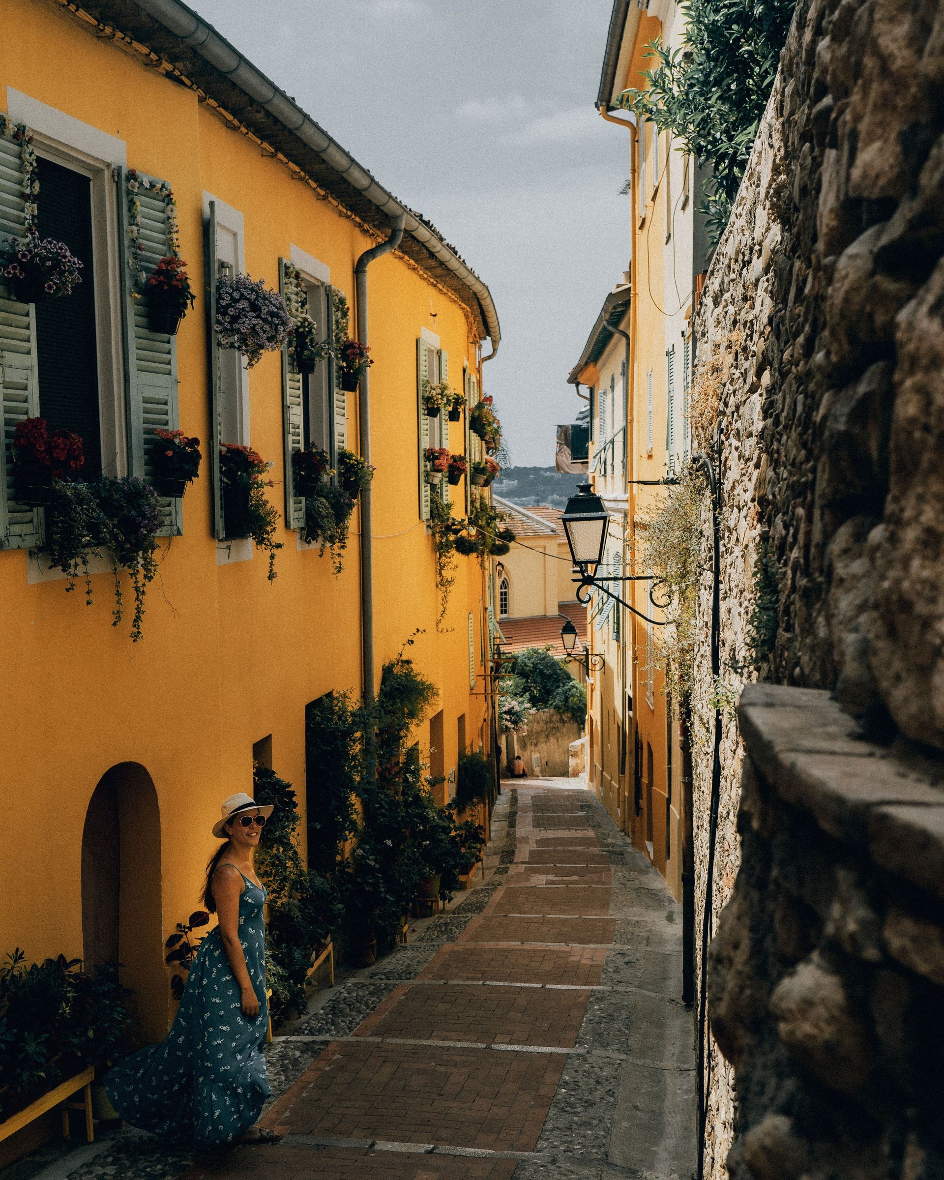 A woman in a blue dress is walking down a narrow alleyway between two yellow buildings
