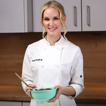 A woman in a chef 's coat is holding a bowl of food and a wooden spoon.