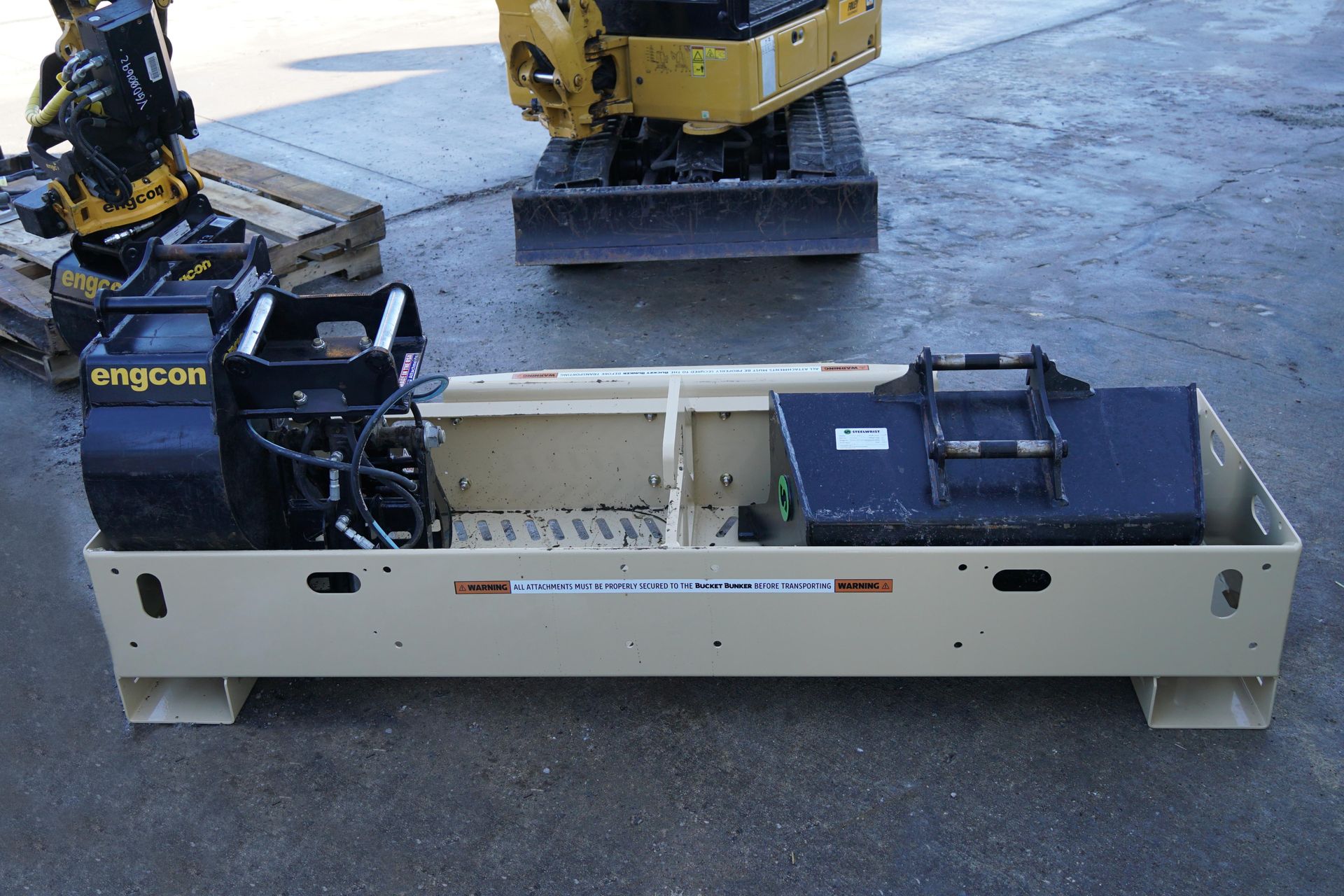 A bucket bunker mini-ex 2t has buckets in it and is sitting Infront of a mini-excavator