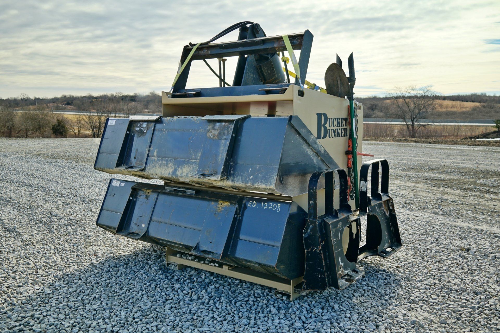 A bucket bunker auger pro is sitting on gravel with attachments loaded on it. 