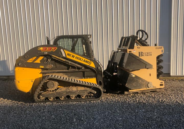 A yellow and black new holland skid loader is parked in front of a building with a bucket bunker and attachments. 