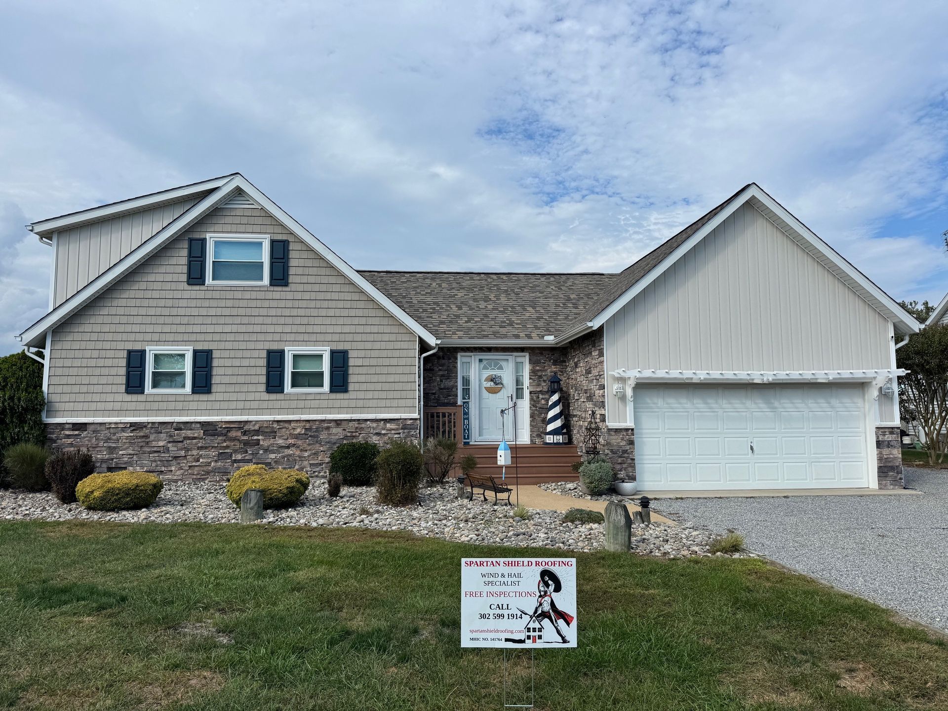House with tan and white siding, stone accents, two-car garage, and a sign in the yard.