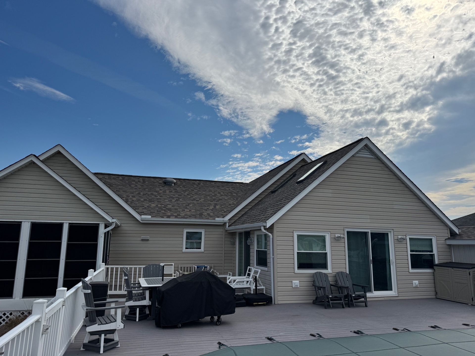 Backyard of a house with a pool and cloudy sky. Beige siding, deck, chairs, grill covered.