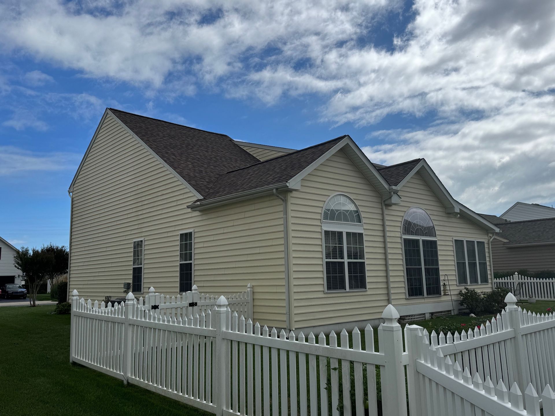 Beige house with brown roof and white picket fence against a cloudy blue sky.