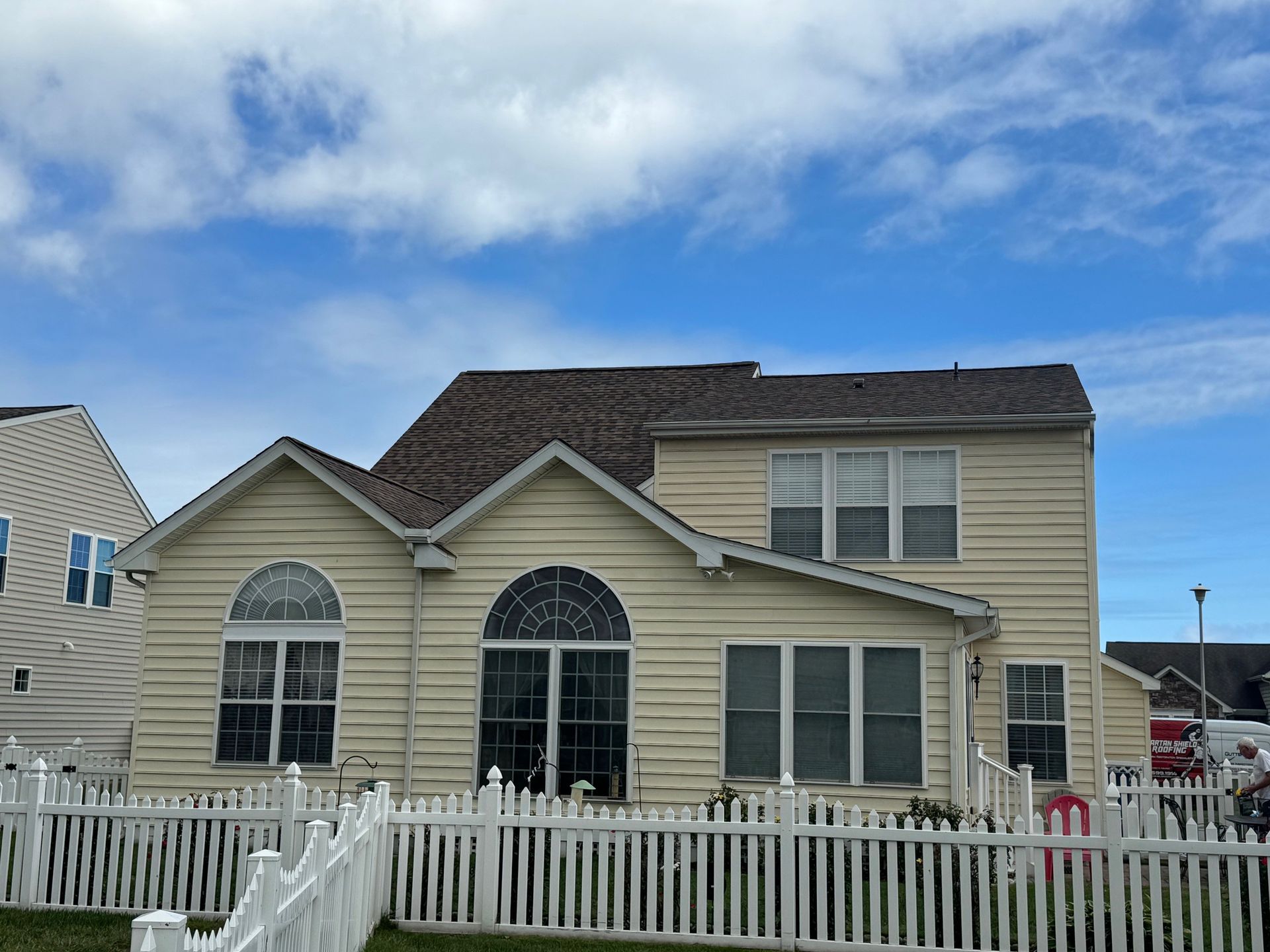 Two-story beige house with a brown roof and a white picket fence under a blue sky.