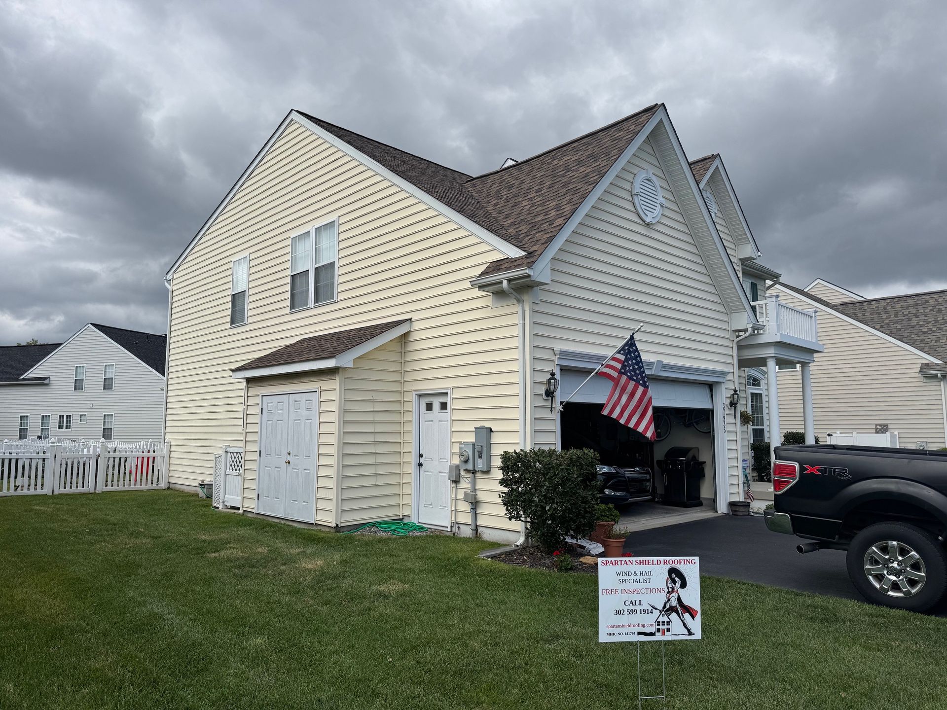 Two-story beige house with a garage and American flag on a cloudy day. Green lawn with a sign in front.