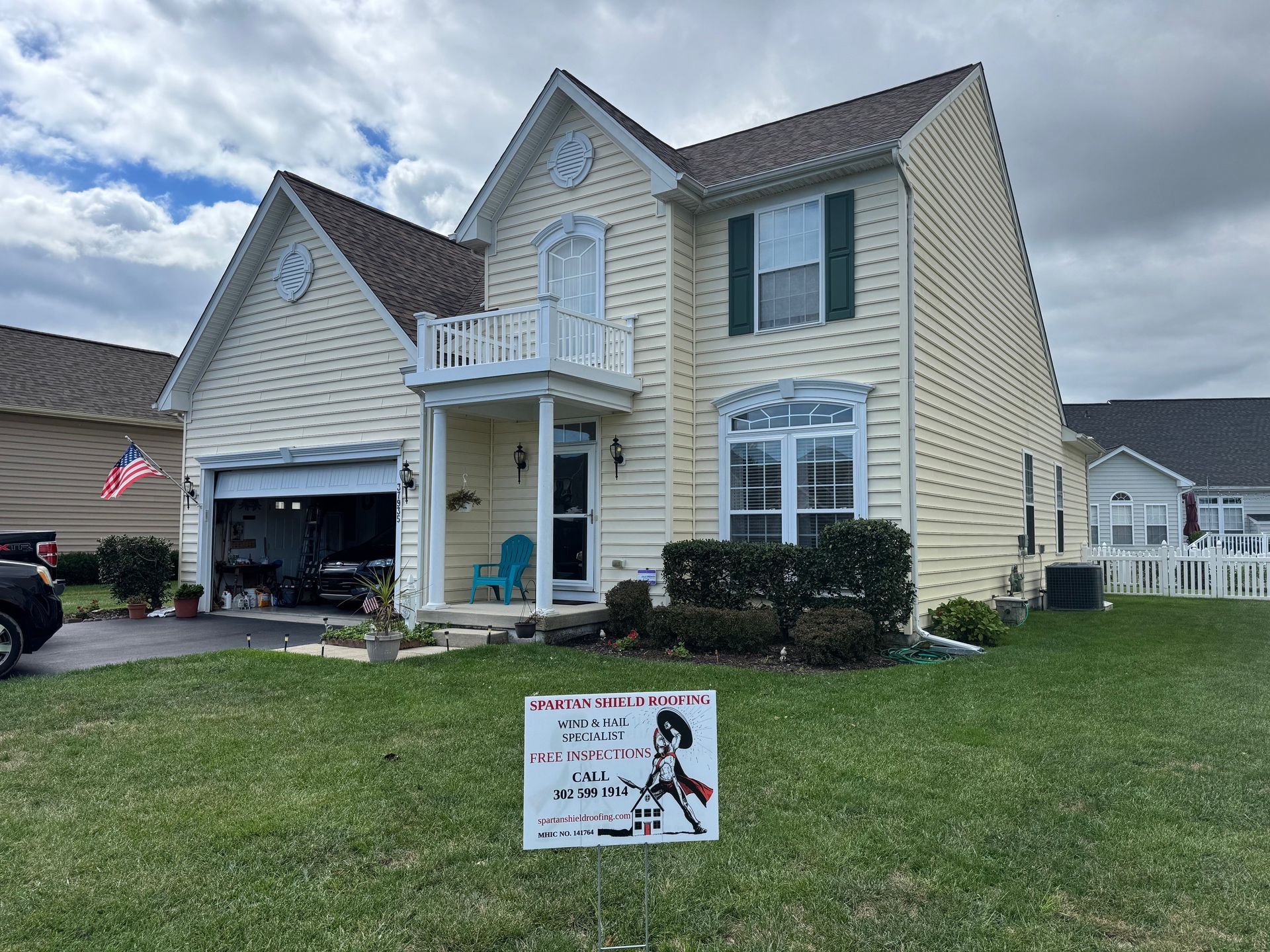 Yellow house with a small balcony, garage open. Sign on lawn. Overcast sky.