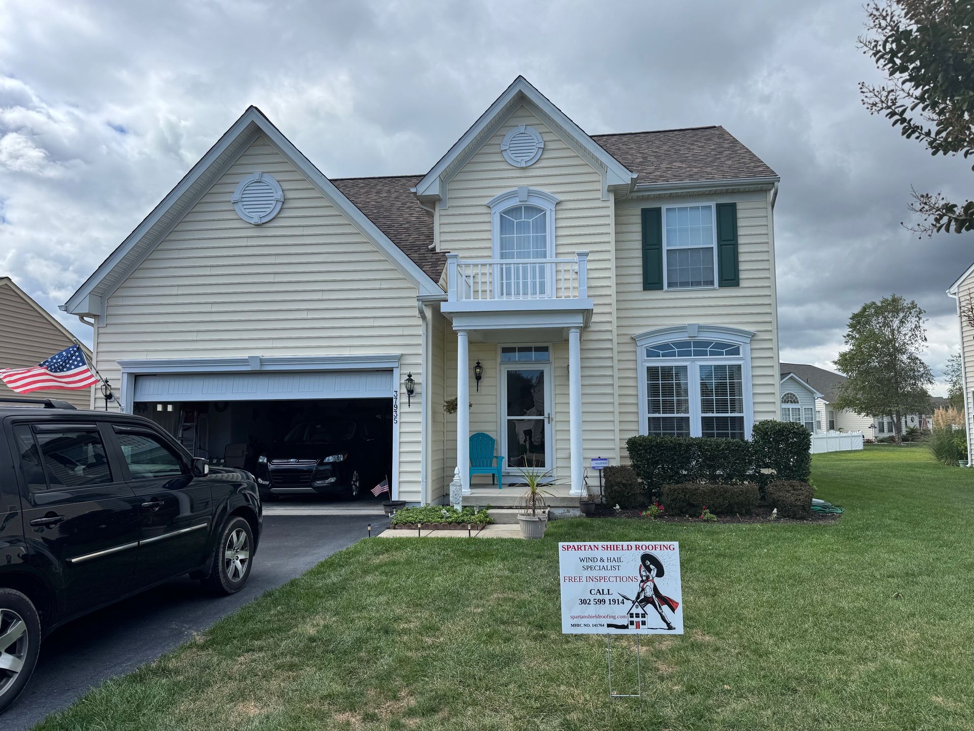 Yellow two-story house with open garage, black SUV, and sign on lawn under cloudy sky.