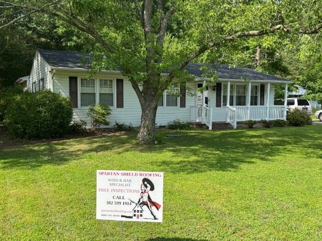 White house with a small porch and a roofing sign in the front yard under a tree on a sunny day.