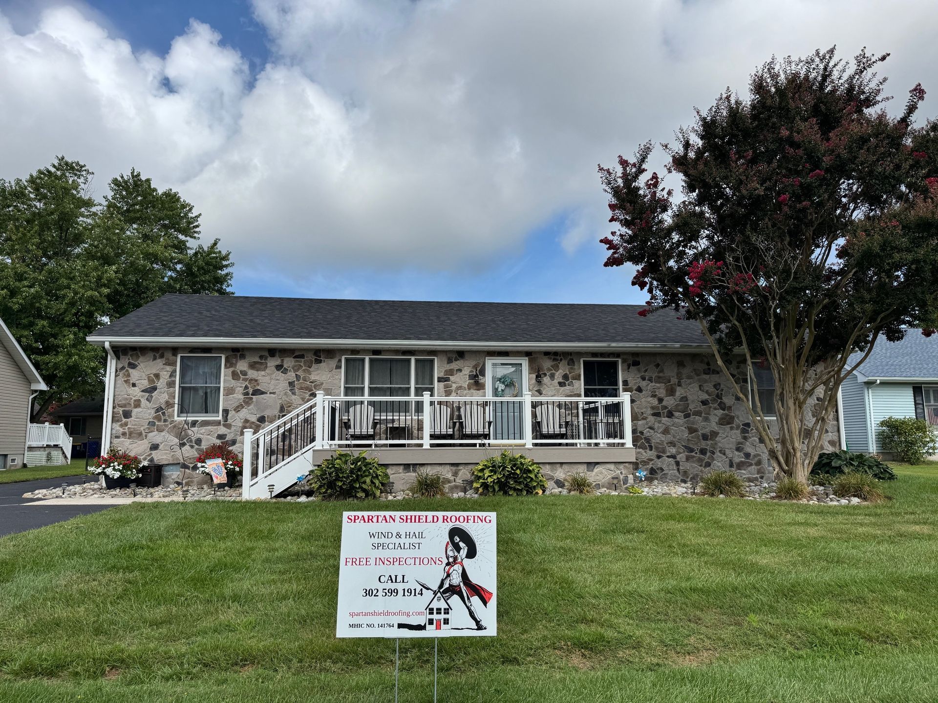 House with stone facade, new dark roof, white porch railing, sign on lawn, cloudy sky.