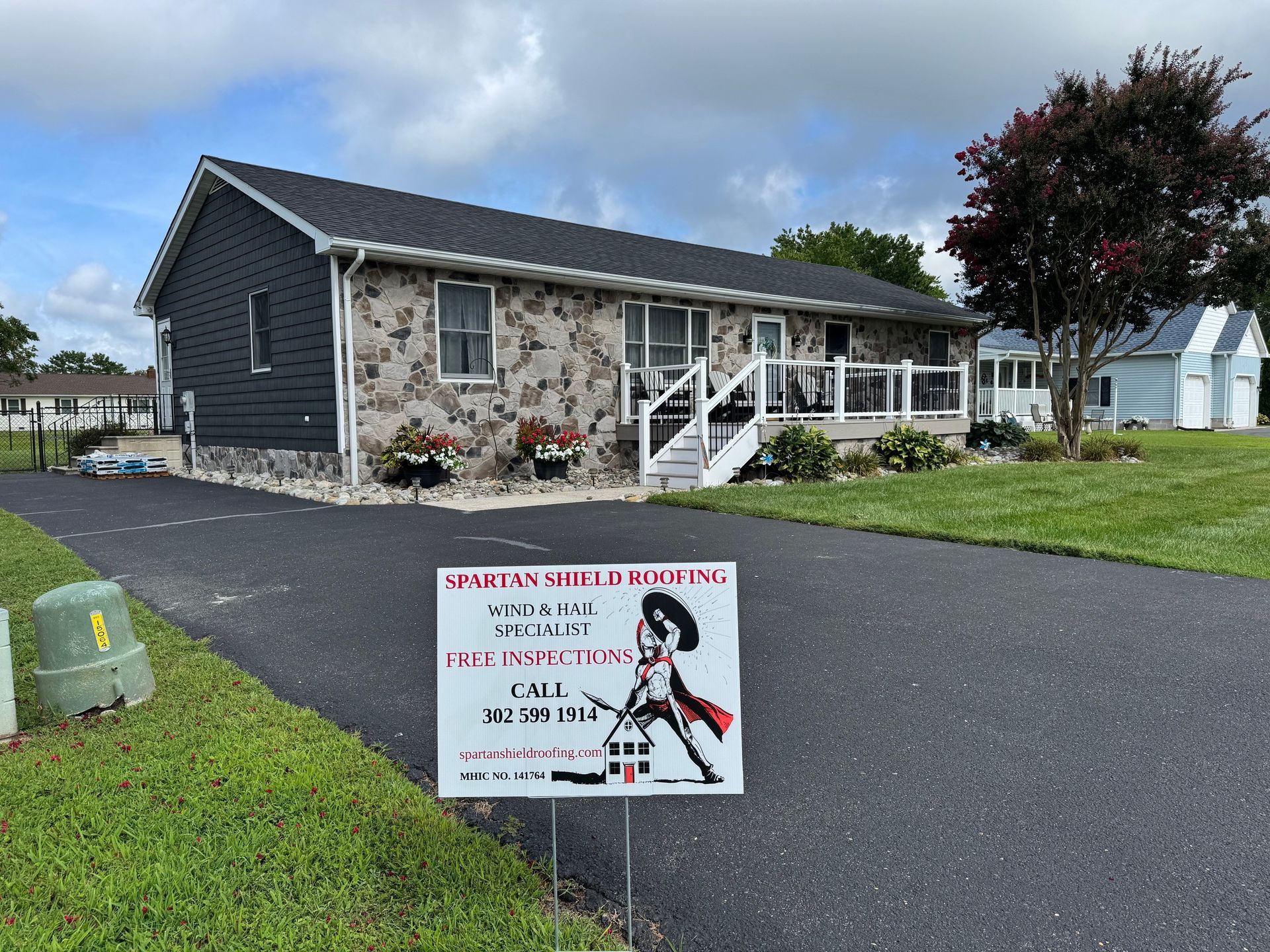 House with stone facade and black siding, sign in driveway advertises roofing services.
