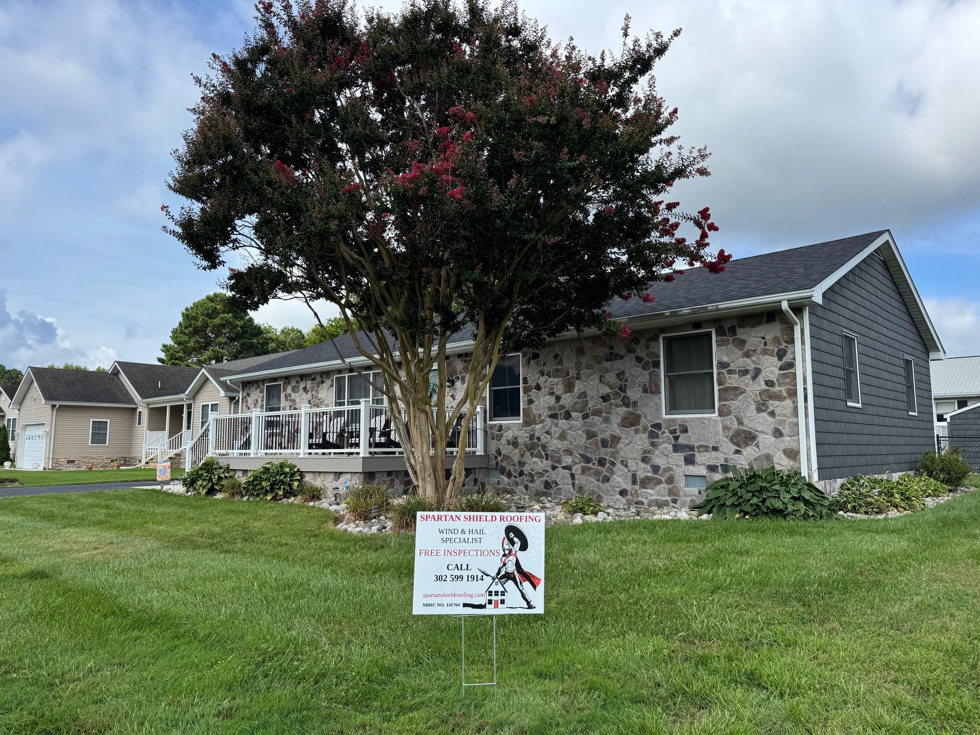 House with stone facade and gray siding, sign in yard under tree.