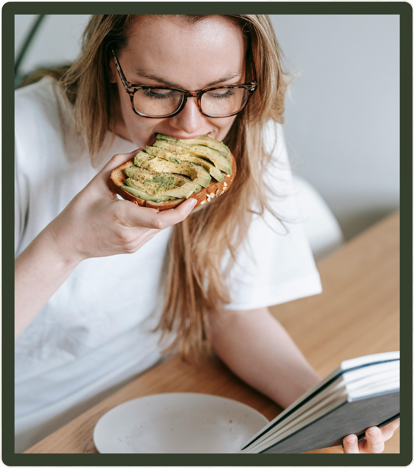 Woman eating avocado toast, reading a book at a table.