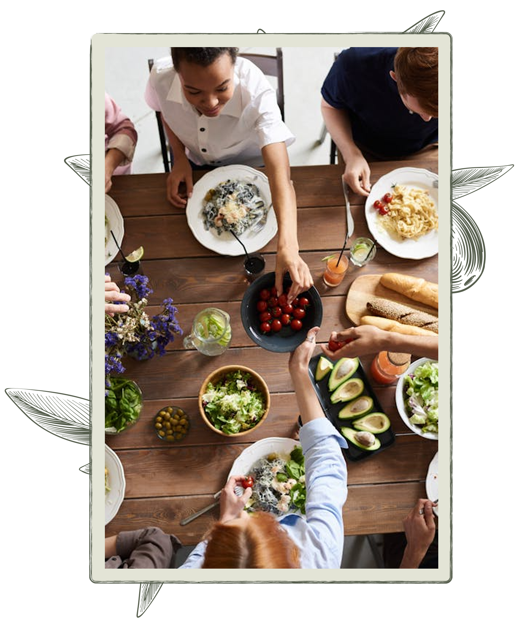 People sharing a meal at a wooden table, passing dishes of food: pasta, salad, avocado, and tomatoes.