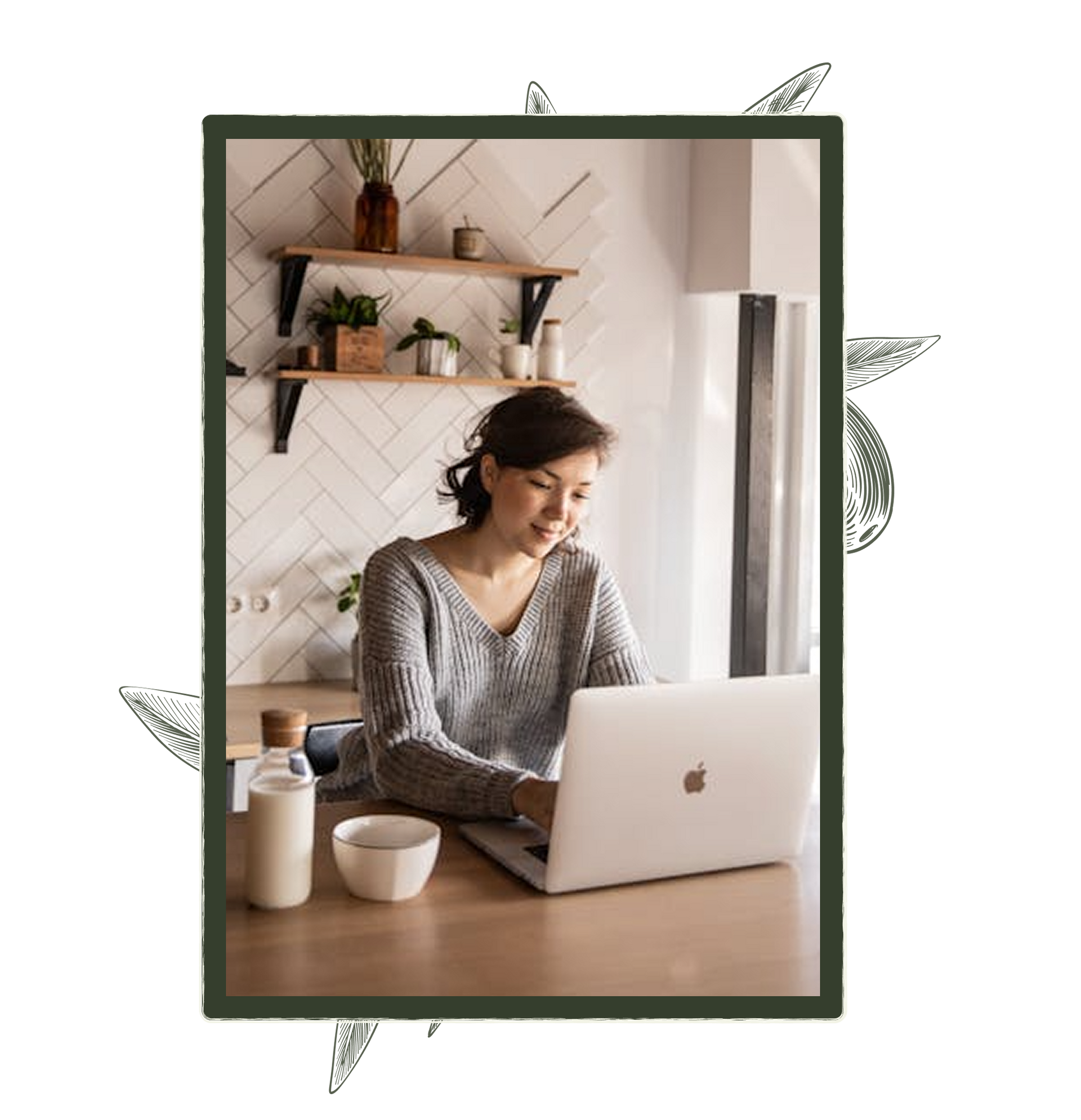 Woman working on a laptop in a kitchen, smiling. A drink and bowl are on the table.