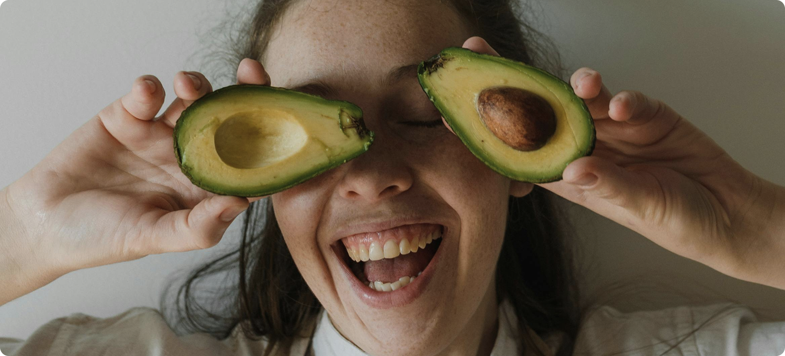 Woman holding avocado halves over eyes, smiling widely.