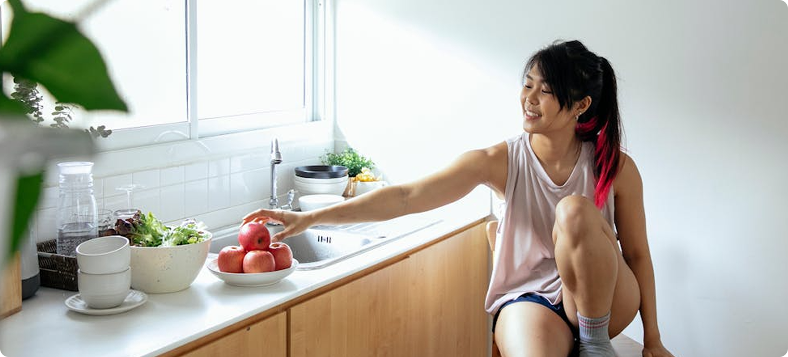 Woman in a kitchen reaching for an apple on a plate by a sink; smiling, natural light.