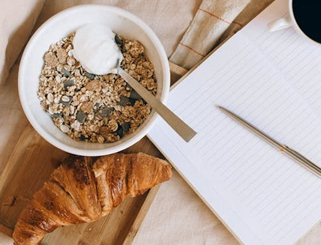 Bowl of cereal with yogurt, croissant, notepad, and coffee cup on a table.