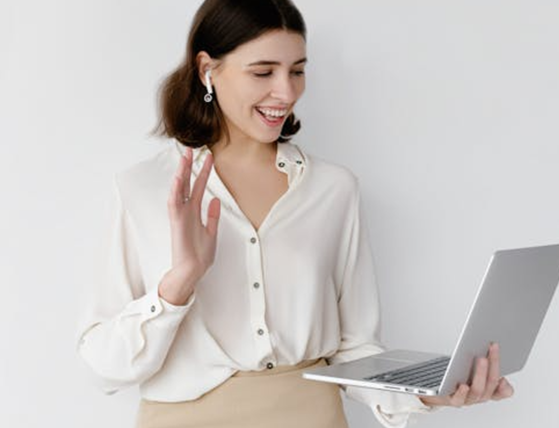 Woman in cream blouse waves at laptop screen, smiles, and wears earbuds.