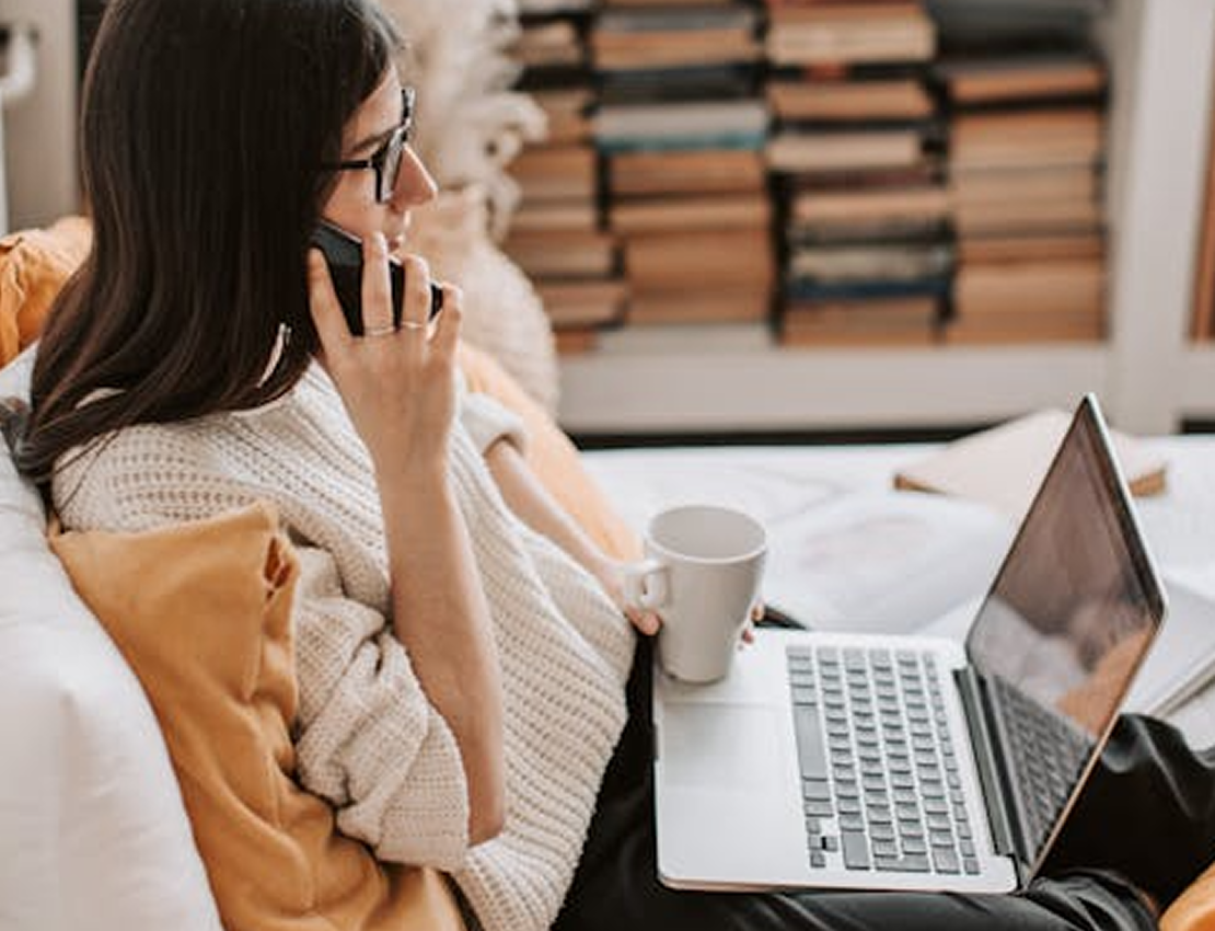 Woman in glasses on the phone, using a laptop, holding a mug, sitting on a couch near bookshelves.
