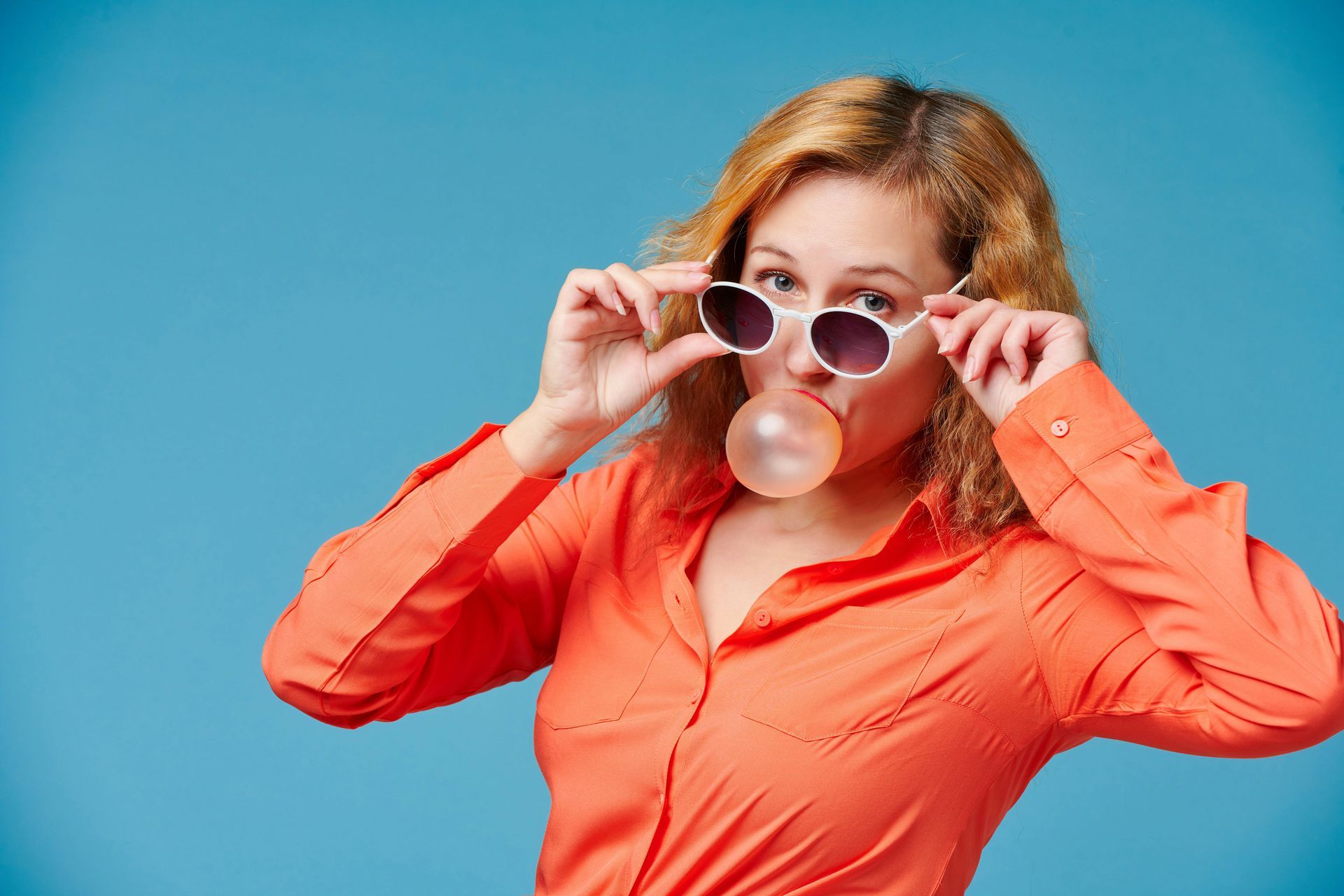 Woman in orange shirt adjusting sunglasses, blowing a pink bubblegum bubble against a blue background