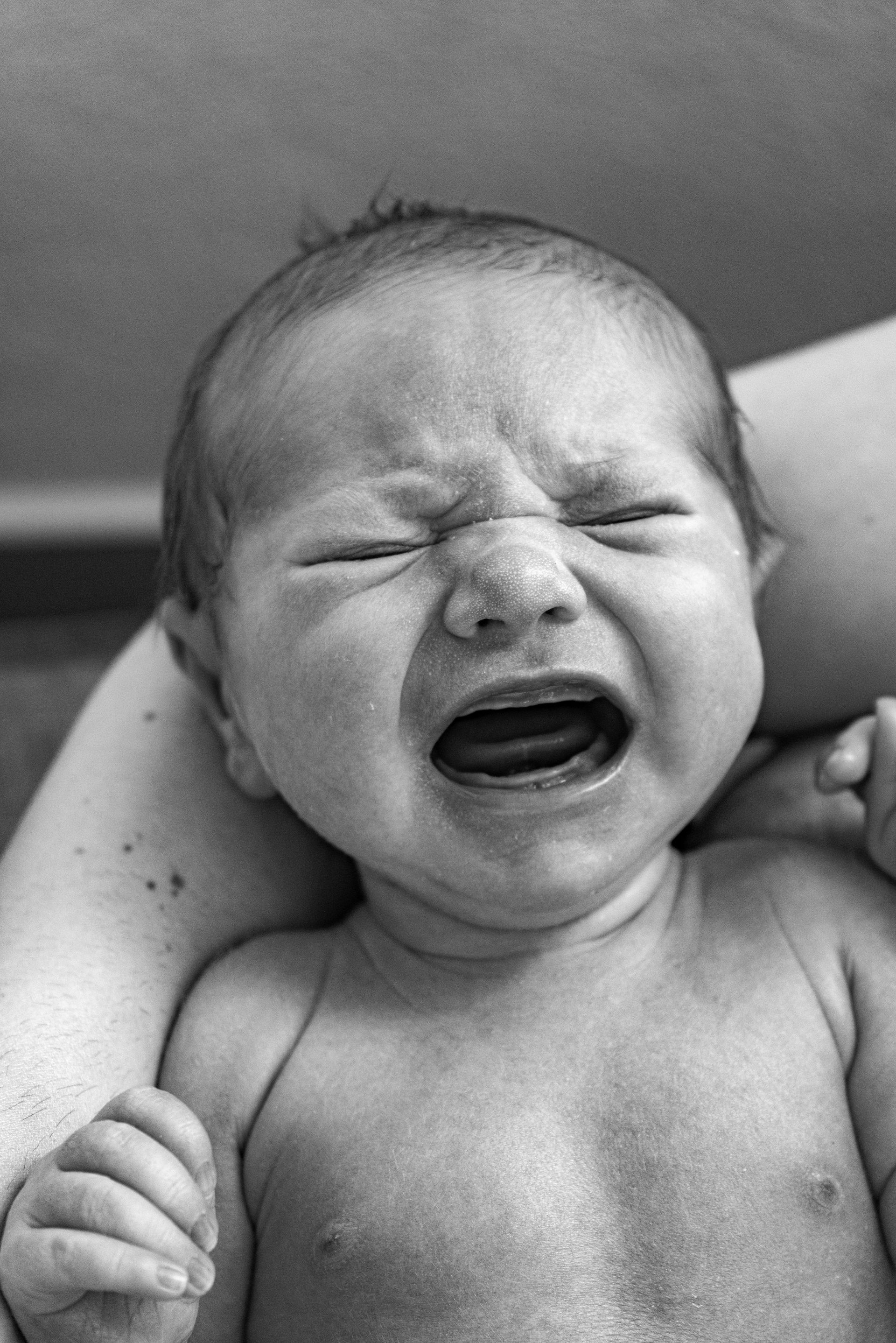 Newborn baby crying, held in someone's arms. Eyes closed, mouth open. Black and white.