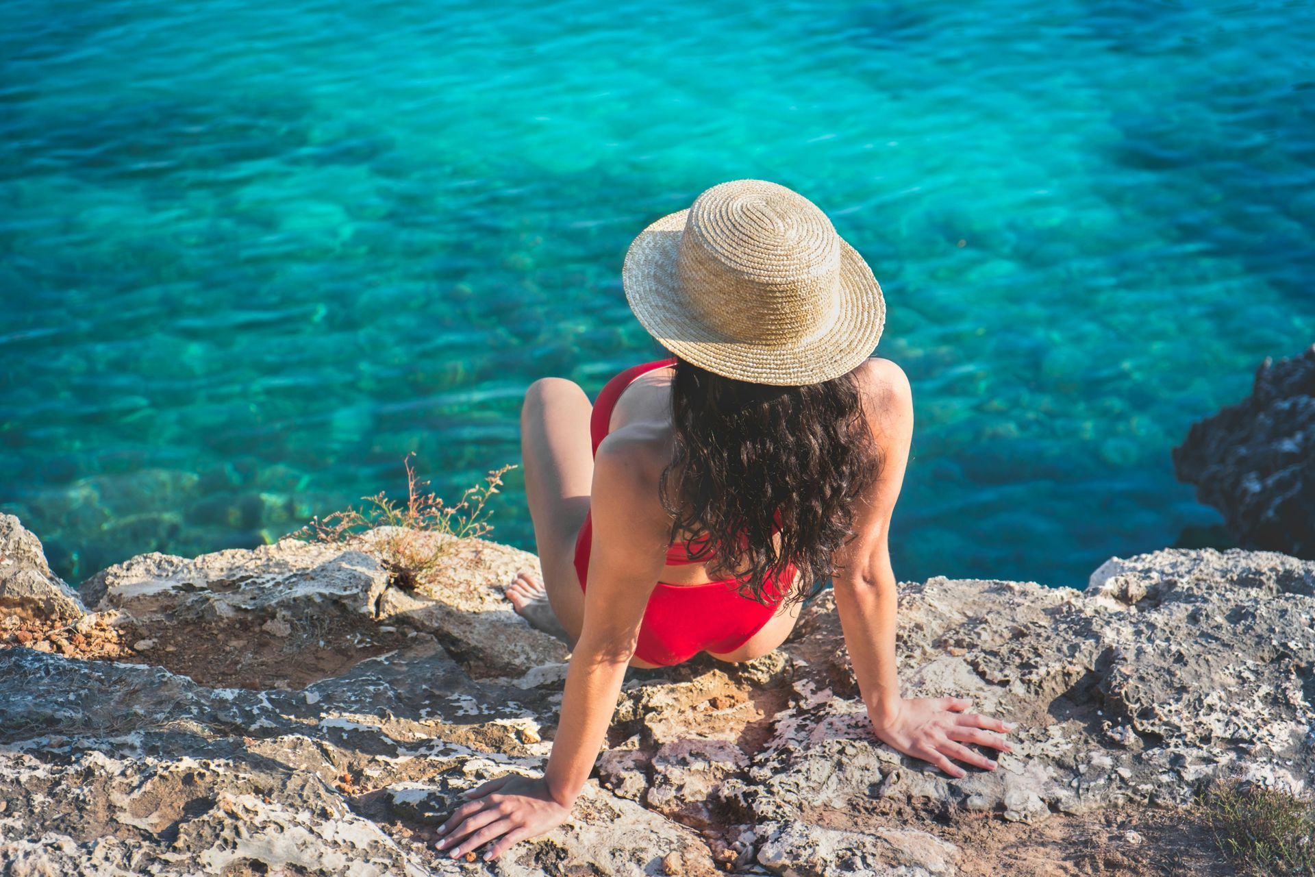 A person in a red swimsuit and straw hat sits on a rocky cliff overlooking vibrant, clear turquoise ocean water.