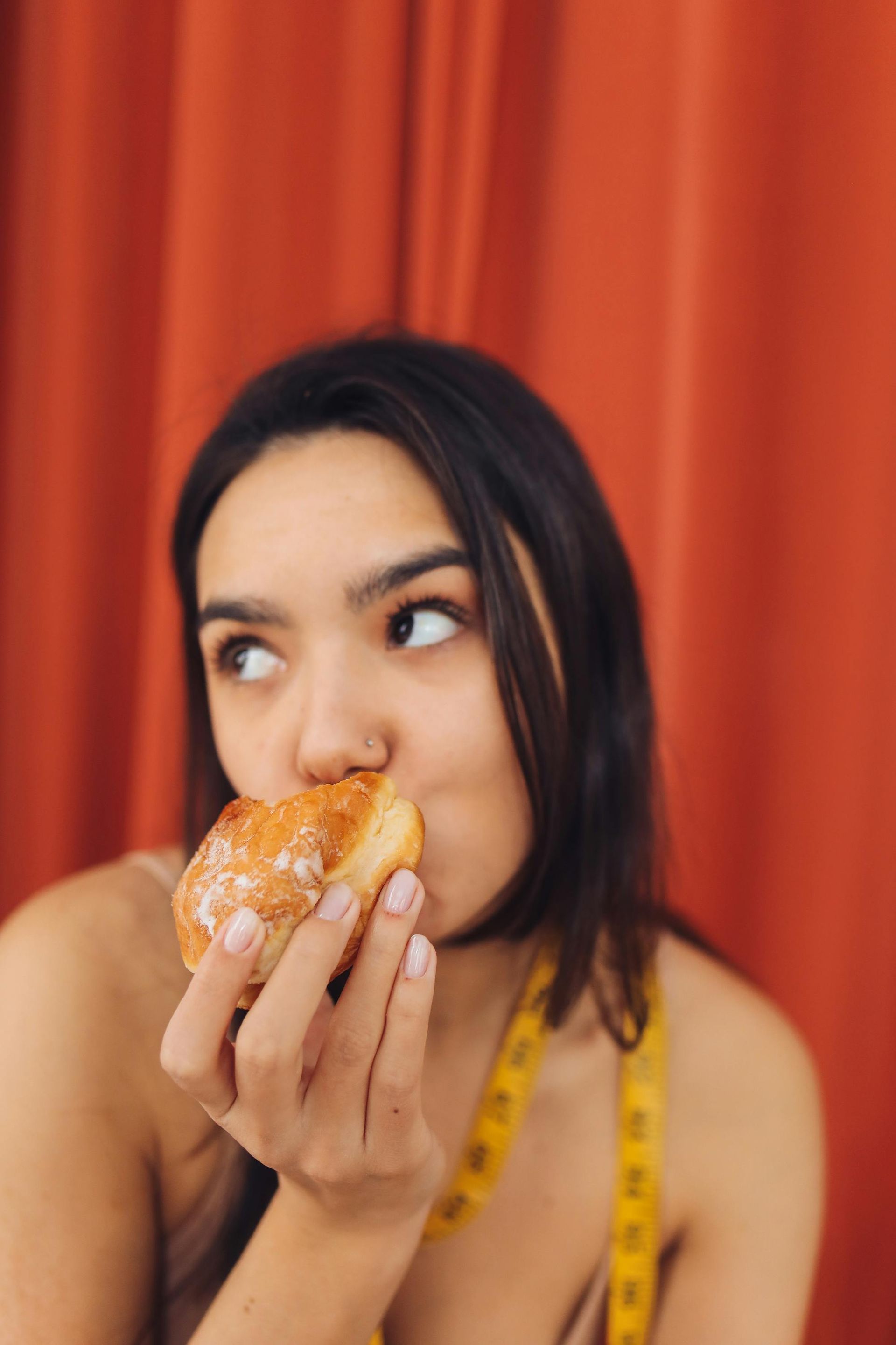 Woman eating a donut, red curtain in the background, measuring tape around her neck, looking to the side.