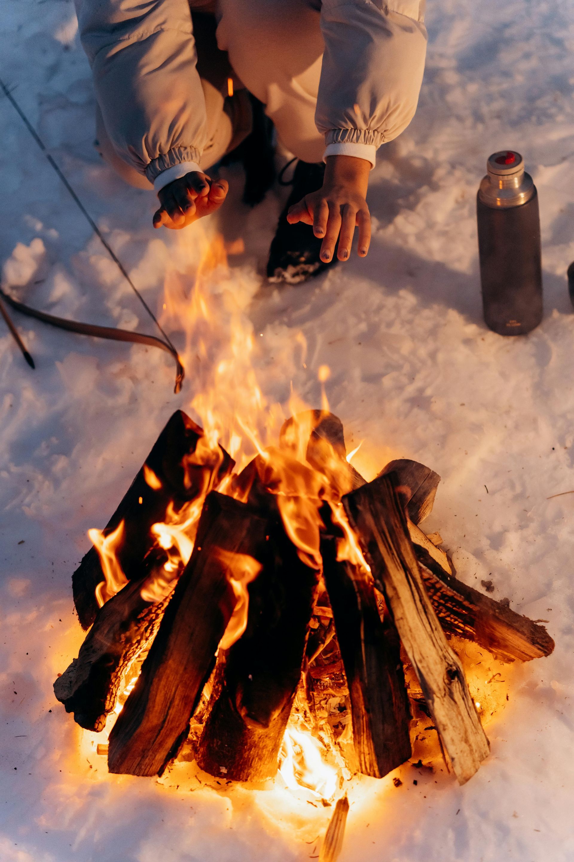 A person near a campfire in the snow. Flames rise from a log pile; a thermos and bow rest nearby.
