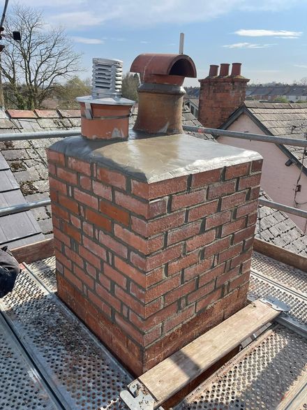 Brick chimney stack on a rooftop with metal flue and surrounding slate roofs under a clear sky