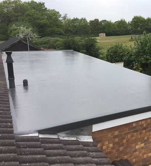 Flat gray roof on a house, with chimney and vent, overlooking trees and a cloudy sky.