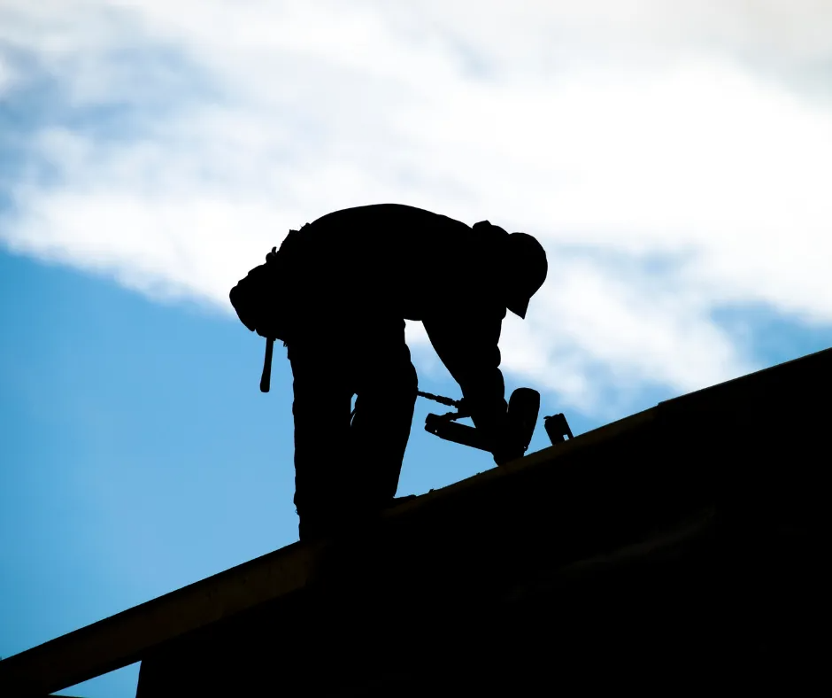Silhouette of a worker on a roof using a hammer against a blue sky