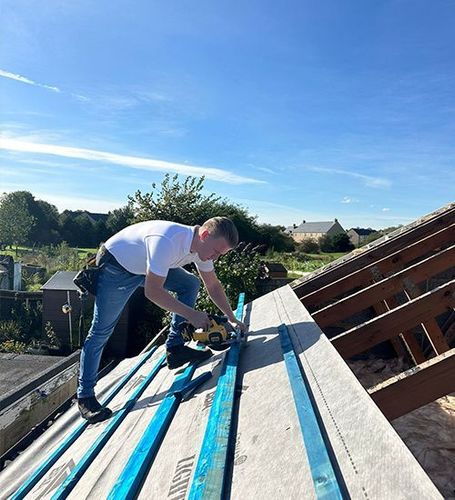Worker using a circular saw to cut roof boards on a rooftop under a clear blue sky