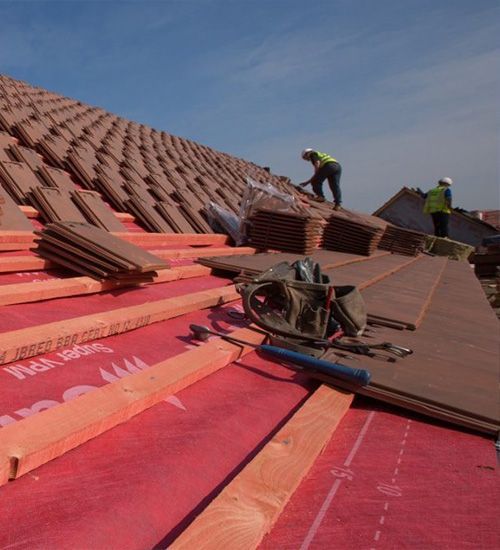 Roofers installing red roofing panels on a steep roof under a blue sky