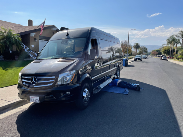 Black van parked on street with person working under it. Sunny day, residential setting.