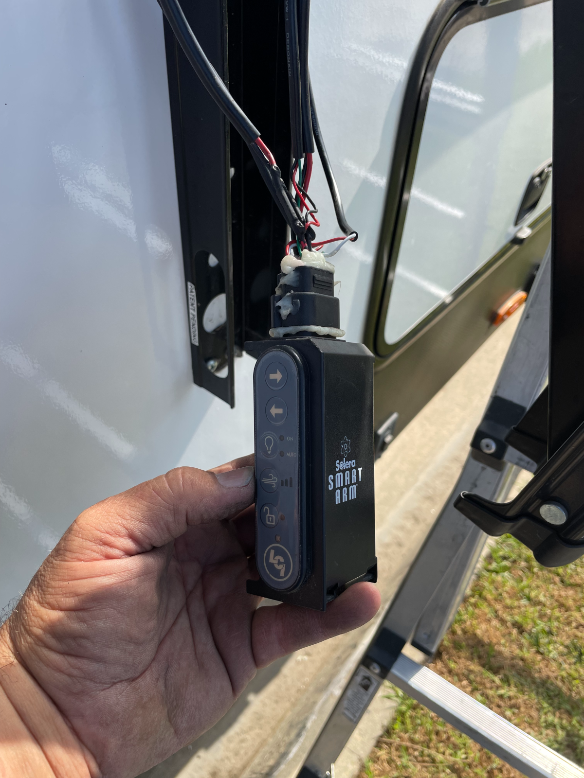 Hand holding a black RV awning control panel with buttons, mounted near a window.