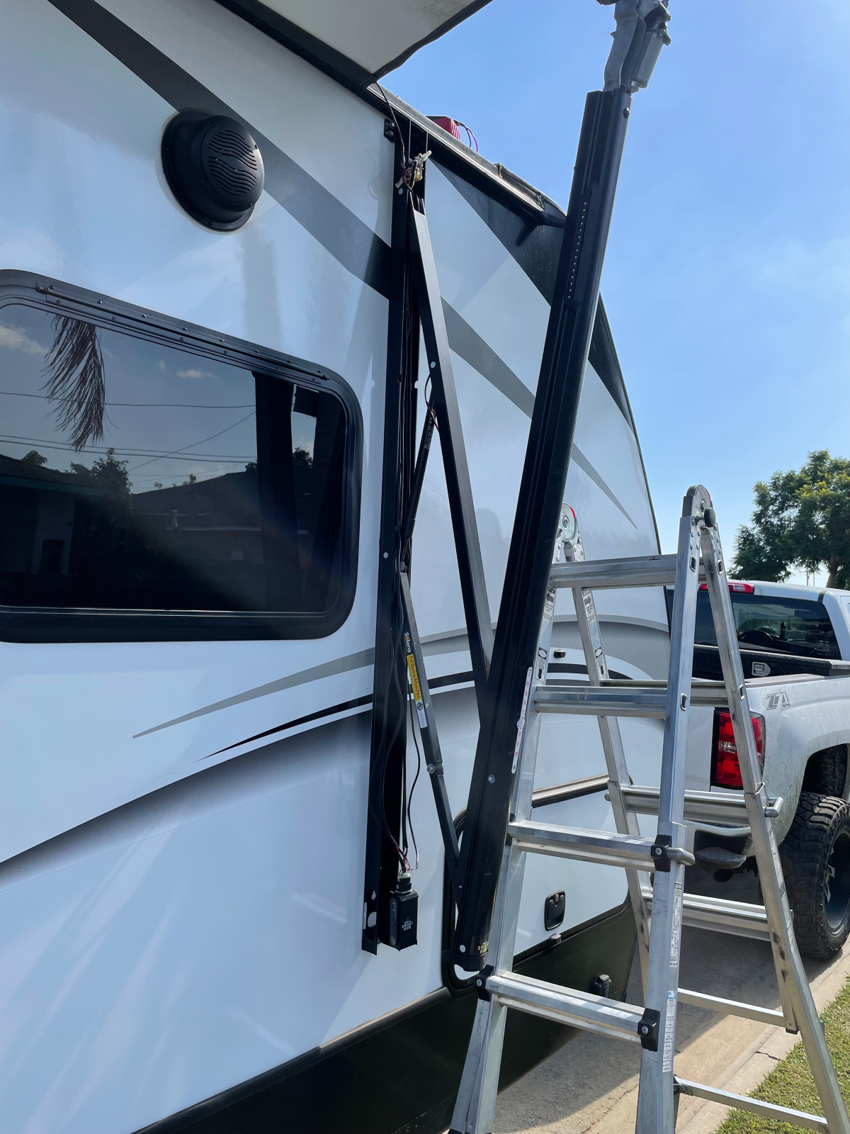 RV side with awning arm extended and a ladder positioned nearby. White and black exterior with blue sky.