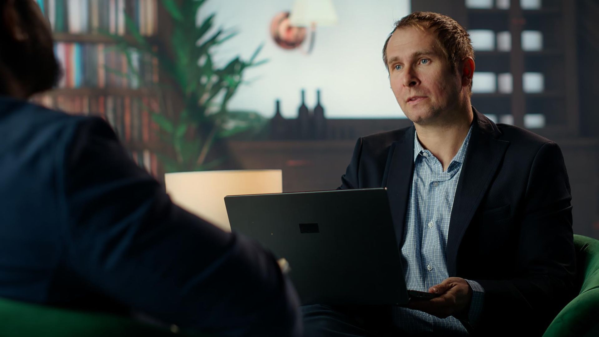 Man in a blazer holds laptop, talking to another person seated in a green armchair. Bookshelf and lamp in background.