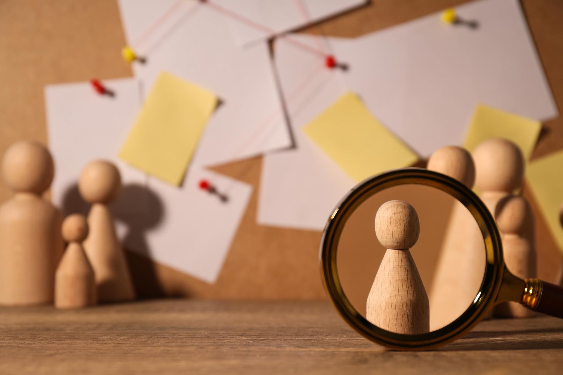 Wooden figurines on a corkboard with notes, one magnified by a hand lens as if lookin.