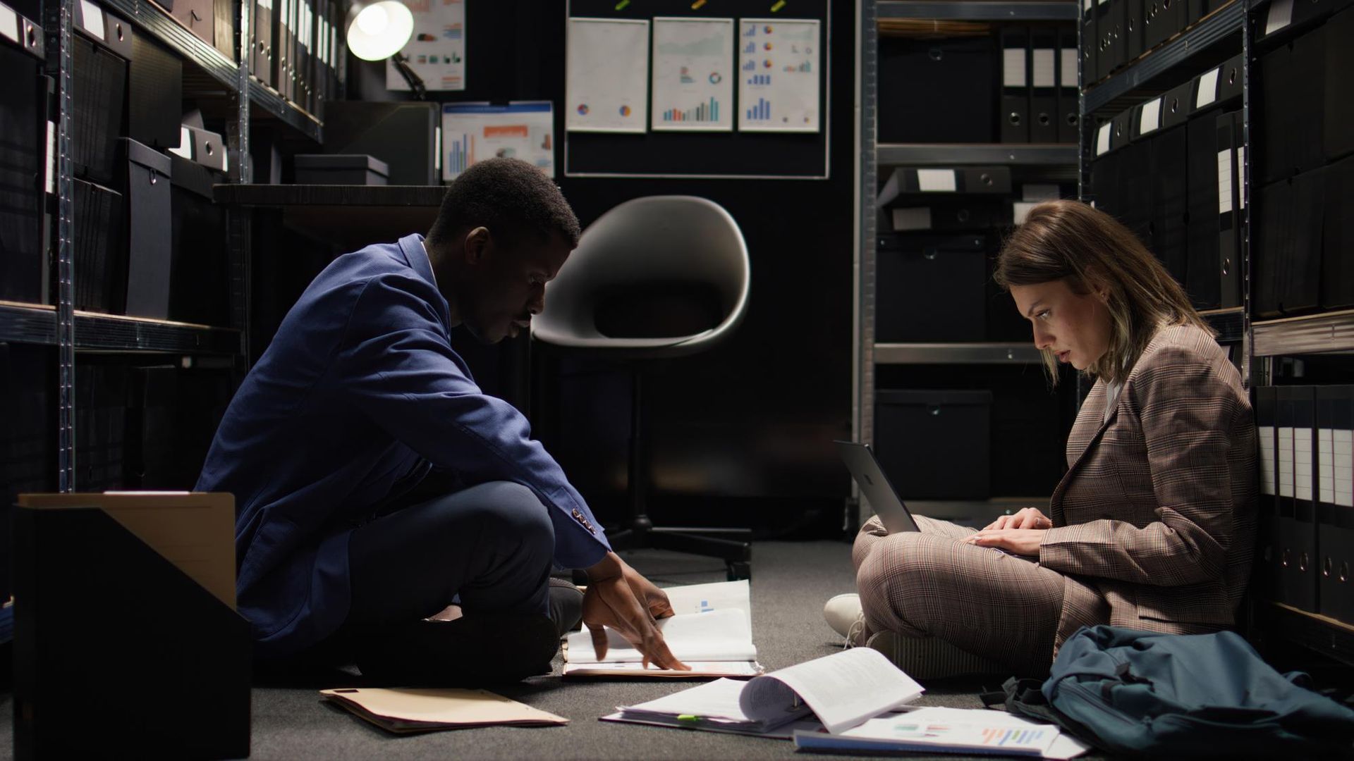 A man and a woman are sitting on the floor in a dark room looking at papers.